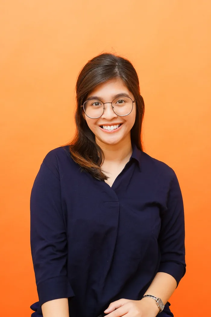 Janine smiling in round glasses and a navy collared top against an orange wall